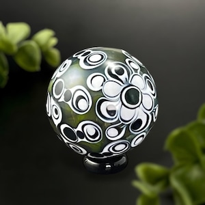May include: A decorative glass orb with a black base, featuring a pattern of white and black floral and circular designs. The orb has a green and black background, and is a unique home decor item.