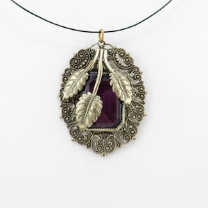 May include: A vintage-style pendant necklace featuring an oval, filigree-style frame in a silver-tone metal. The pendant has a rectangular, deep purple gemstone at its center, with gold-colored leaf accents.