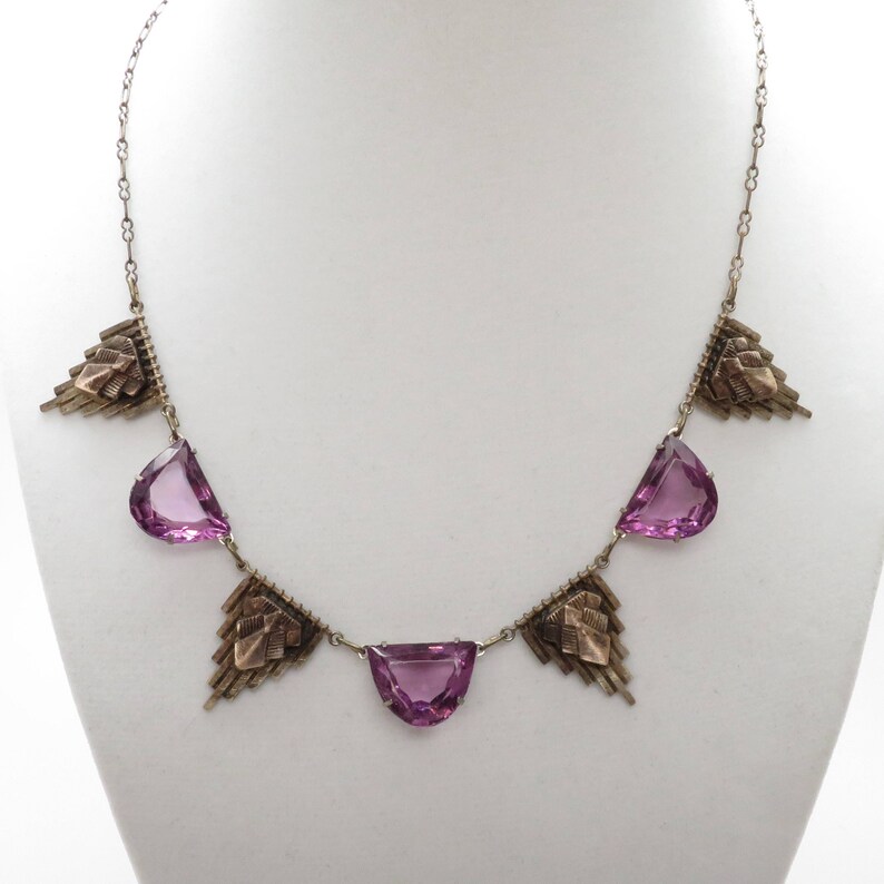 K&ouml;nnte beinhalten: Eine silberne Halskette mit geometrischem Design verf&uuml;gt &uuml;ber f&uuml;nf facettierte violette Steine und goldene Akzente.