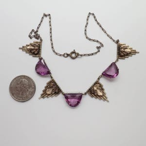 K&ouml;nnte beinhalten: Eine silberne Halskette mit geometrischem Design und violetten Steinen. Die Halskette besteht aus einer silbernen Kette und hat ein geometrisches Design mit violetten Steinen. Die Halskette ist etwa 46 cm lang.