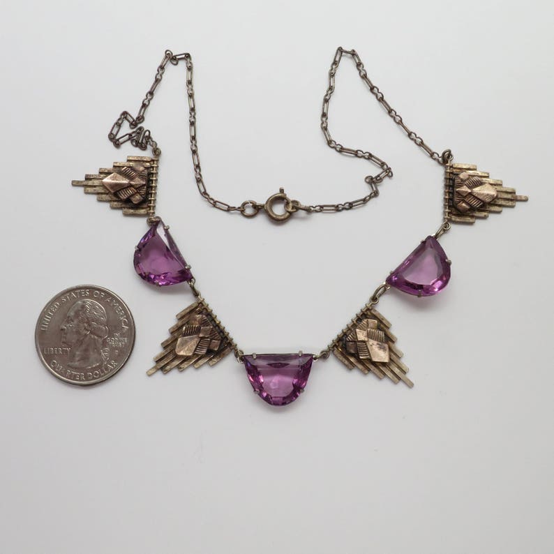 K&ouml;nnte beinhalten: Eine silberne Halskette mit geometrischem Design und violetten Steinen. Die Halskette besteht aus einer silbernen Kette und hat ein geometrisches Design mit violetten Steinen. Die Halskette ist etwa 46 cm lang.
