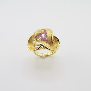 以下が含まれることがあります： 中央にピンクの宝石がセットされた、テクスチャ加工のデザインの金のリングです。