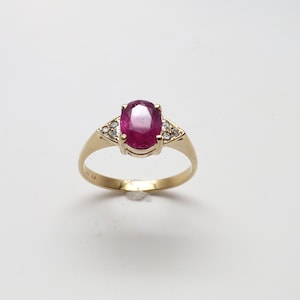 May include: A gold ring with a large oval-cut ruby gemstone. The ruby is a deep red hue. Small, clear gemstones are set on either side of the ruby. The ring has a classic design. The ring is a piece of jewellery.