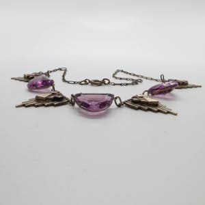 K&ouml;nnte beinhalten: Eine silberne Halskette mit geometrischem Design und drei gro&szlig;en, facettierten, violetten Steinen.