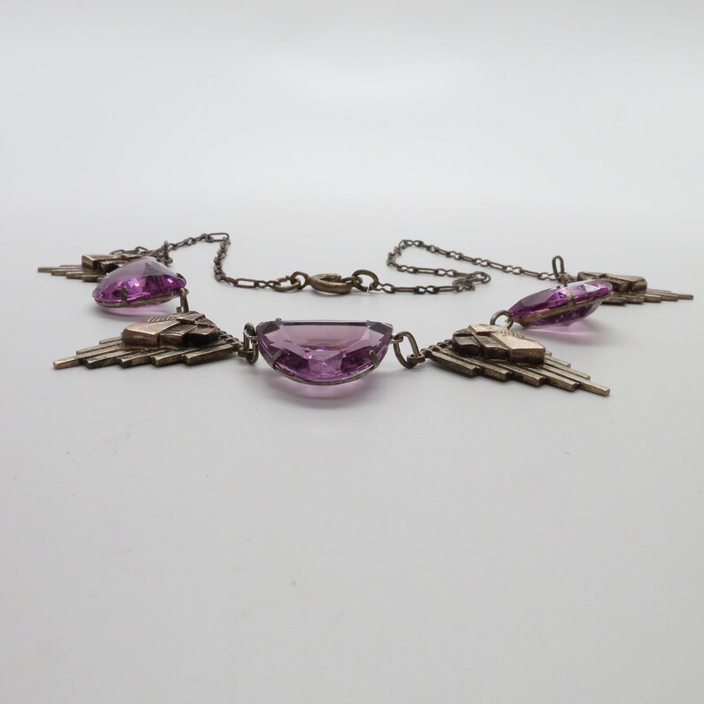 K&ouml;nnte beinhalten: Eine silberne Halskette mit geometrischem Design und drei gro&szlig;en, facettierten, violetten Steinen.