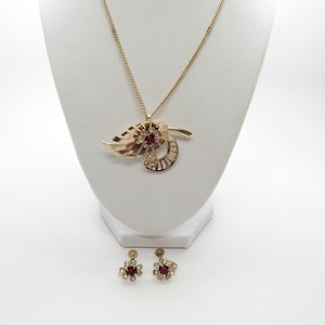 May include: A gold-coloured necklace and earring set. The necklace has a pendant with a leaf design, a central red stone, and clear accent stones. The matching earrings are small, flower-shaped, with red and clear stones. The jewellery is displayed on a white bust.
