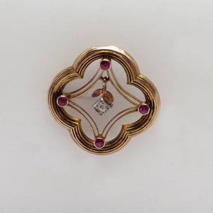 May include: A gold brooch with a clear center and four red gemstones. A small diamond hangs from the center.