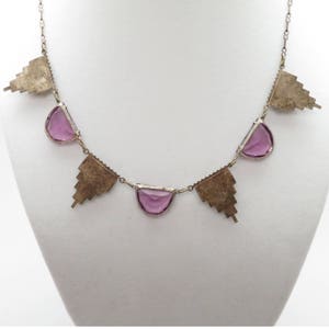 K&ouml;nnte beinhalten: Eine silberne Halskette mit geometrischem Design und violetten Glassteinen. Die Halskette hat eine zarte Kette und zeigt ein sich wiederholendes Muster aus Dreiecken und Halbkreisen.