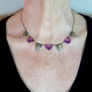K&ouml;nnte beinhalten: Eine silberne Halskette mit einer zarten Kette und f&uuml;nf violetten Edelsteinanh&auml;ngern. Jeder Anh&auml;nger ist tropfenf&ouml;rmig und von einem silbernen Design umgeben, das an Fl&uuml;gel erinnert.