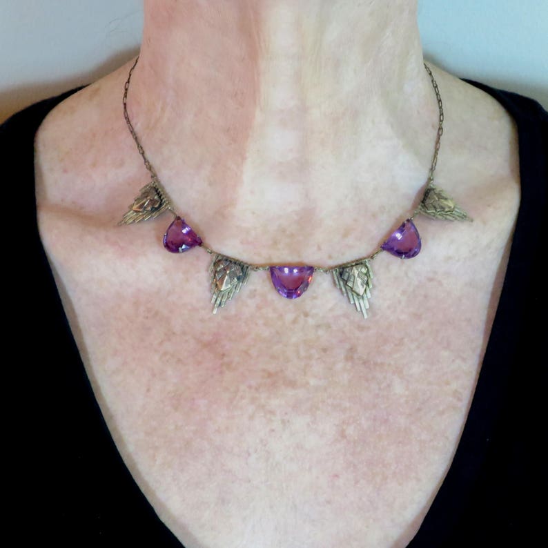 K&ouml;nnte beinhalten: Eine silberne Halskette mit einer zarten Kette und f&uuml;nf violetten Edelsteinanh&auml;ngern. Jeder Anh&auml;nger ist tropfenf&ouml;rmig und von einem silbernen Design umgeben, das an Fl&uuml;gel erinnert.
