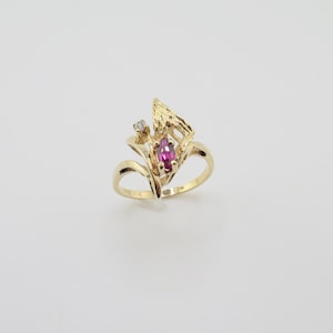 May include: A gold ring with a pink oval gemstone and a small diamond accent. The ring has a unique, abstract design.