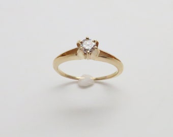 Könnte beinhalten: Ein goldener Ring mit einem einzigen, klaren, runden Edelstein, der in einer sechsarmigen Fassung sitzt.