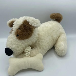 May include: A plush dog toy with a cream-colored body, brown patches around the eye and ear, and a brown tail. The dog is holding a cream-colored bone-shaped toy. The dog's nose is black.