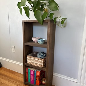 May include: A dark brown wooden bookshelf with three shelves. The top shelf has two small bowls, one blue and white and the other pink and white. The middle shelf has a woven basket with a blue and white patterned fabric inside. The bottom shelf has a row of colorful books and a small orange bowl.