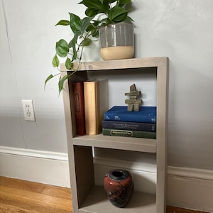 May include: A gray wooden shelf unit with three shelves. The top shelf has a potted plant with green leaves. The middle shelf has a stack of books and a small wooden sculpture. The bottom shelf has a red and black ceramic vase.