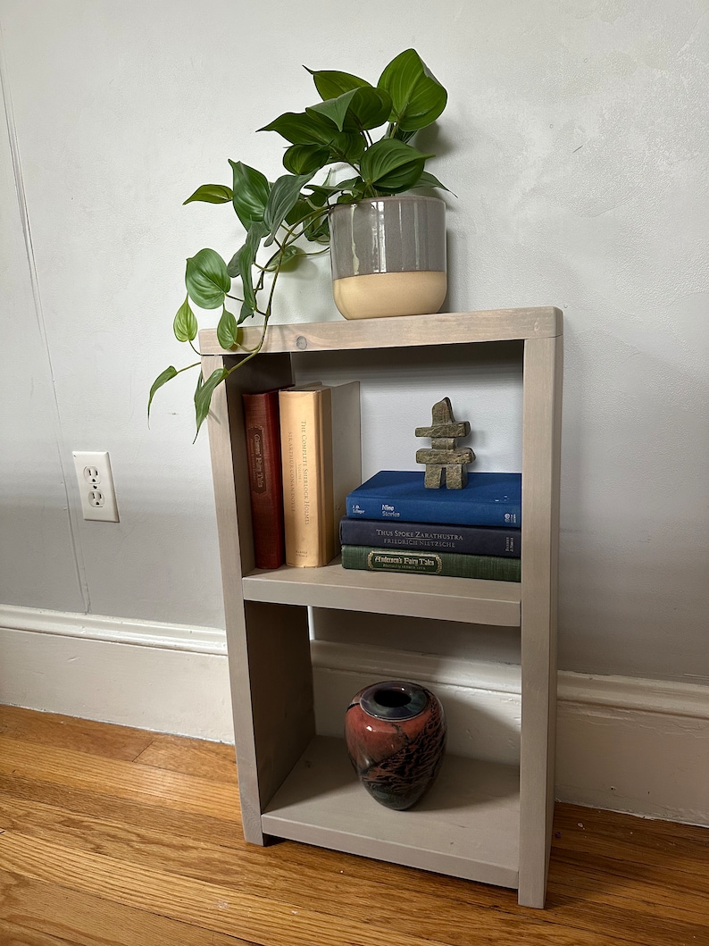 May include: A gray wooden shelf unit with three shelves. The top shelf has a potted plant with green leaves. The middle shelf has a stack of books and a small wooden sculpture. The bottom shelf has a red and black ceramic vase.