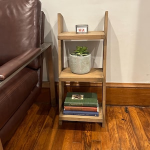 May include: A wooden three-tiered shelf unit with a small potted succulent plant, a candle with the word "HA" on it, and a stack of three books on the bottom shelf.