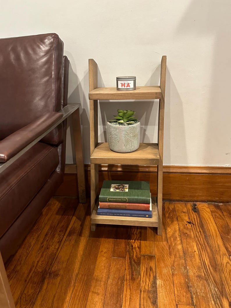 May include: A wooden three-tiered shelf unit with a small potted succulent plant, a candle with the word "HA" on it, and a stack of three books on the bottom shelf.