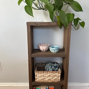 May include: A dark brown wooden shelf unit with three shelves. The top shelf has two small bowls, one pink and one blue. The middle shelf has a woven basket with a blue cloth inside. The bottom shelf has a stack of six books with colorful covers. The books have the title "A Court of Thorns and Roses" and the author "Sarah J. Maas" on the spines.