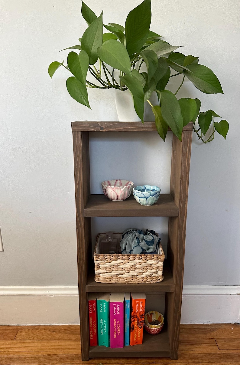 May include: A dark brown wooden shelf unit with three shelves. The top shelf has two small bowls, one pink and one blue. The middle shelf has a woven basket with a blue cloth inside. The bottom shelf has a stack of six books with colorful covers. The books have the title "A Court of Thorns and Roses" and the author "Sarah J. Maas" on the spines.