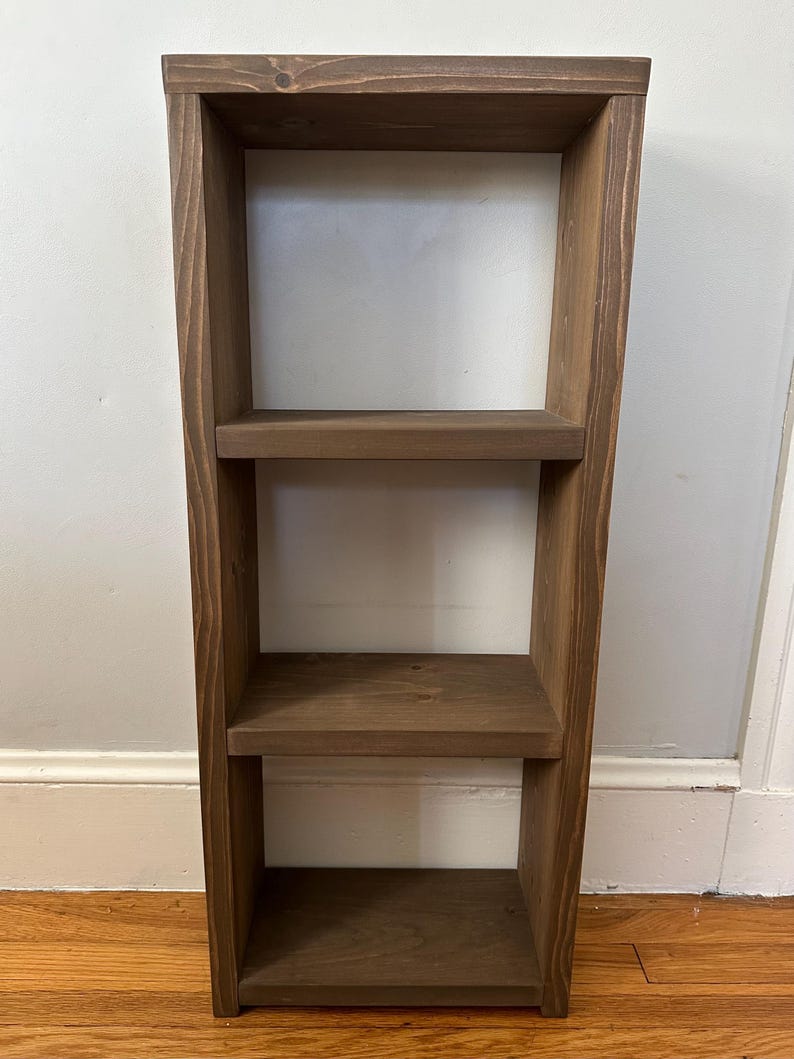 May include: A three-tiered wooden shelf unit with a dark brown stain. The shelf is made of solid wood and has a simple, rustic design.