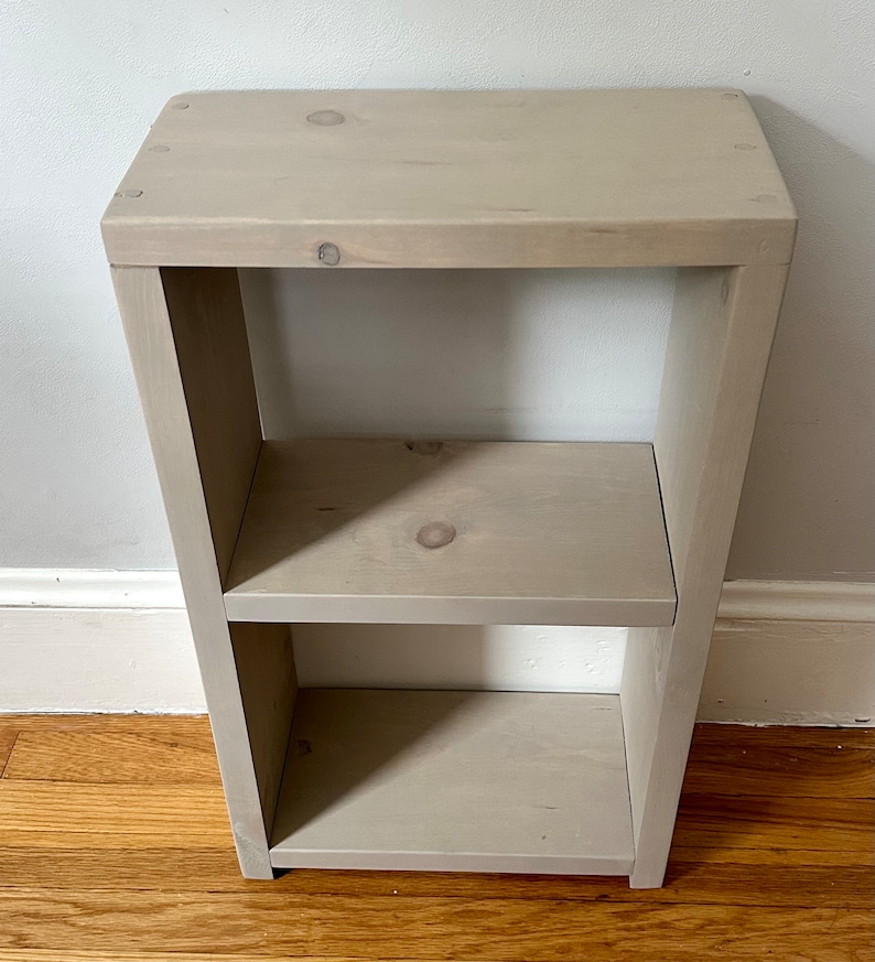 May include: A light gray wooden shelf unit with two shelves. The shelf unit is made of unfinished wood and has a simple, modern design.