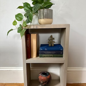 May include: A white wooden shelf unit with two shelves. The top shelf has a potted plant, a wooden sculpture, and three books. The bottom shelf has a decorative vase.