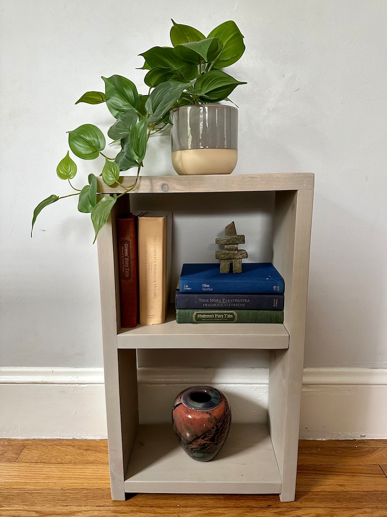 May include: A white wooden shelf unit with two shelves. The top shelf has a potted plant, a wooden sculpture, and three books. The bottom shelf has a decorative vase.
