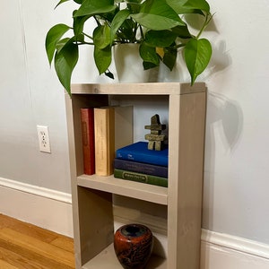 May include: A light gray wooden bookshelf with two shelves. The top shelf has three books and a small stone sculpture. The bottom shelf has a small, red and black ceramic vase. A potted plant sits on top of the bookshelf.