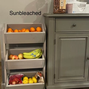 May include: A three-tiered wooden fruit and vegetable stand with a gray finish. The stand is filled with oranges, bananas, apples, onions, and lemons. The stand is next to a gray cabinet with a drawer and a door. The text "Sunbleached" is written on the wall behind the stand.