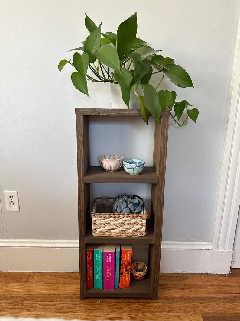 May include: A wooden shelf unit with four shelves, featuring a potted plant, two small bowls, a woven basket, and a stack of books with the title "A Court of Thorns and Roses" on the bottom shelf.