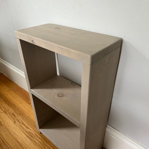May include: A small, light gray wooden shelf unit with two shelves. The shelf unit is made of unfinished wood and has a rustic look.