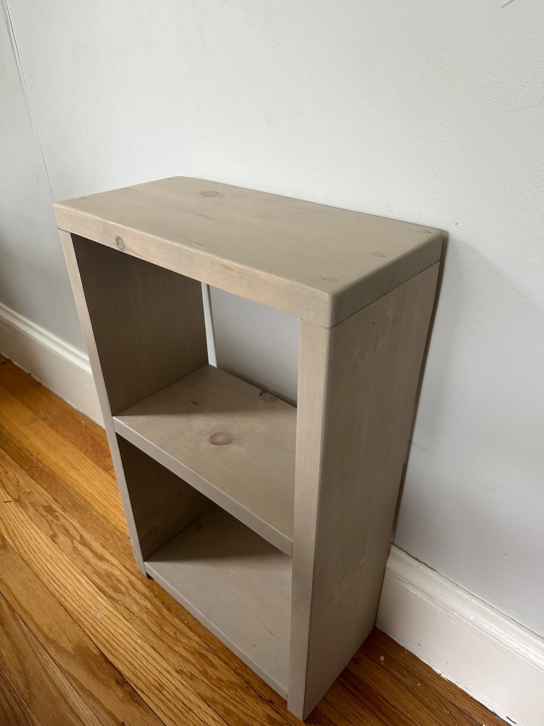 May include: A small, light gray wooden shelf unit with two shelves. The shelf unit is made of unfinished wood and has a rustic look.