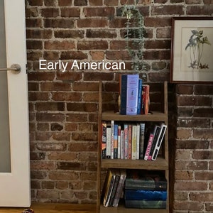 K&ouml;nnte beinhalten: Ein B&uuml;cherregal aus Holz in Form einer Leiter mit mehreren Regalbrettern, die mit B&uuml;chern gef&uuml;llt sind. Das B&uuml;cherregal steht an einer Backsteinwand mit dem Text "Early American" an der Wand.