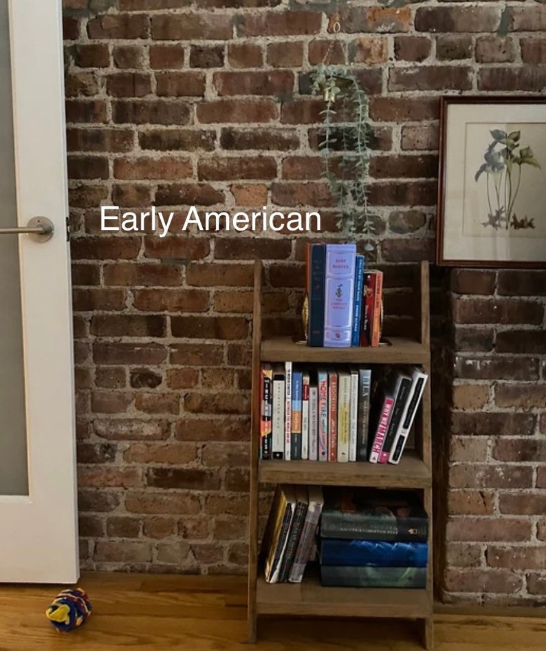 K&ouml;nnte beinhalten: Ein B&uuml;cherregal aus Holz in Form einer Leiter mit mehreren Regalbrettern, die mit B&uuml;chern gef&uuml;llt sind. Das B&uuml;cherregal steht an einer Backsteinwand mit dem Text "Early American" an der Wand.