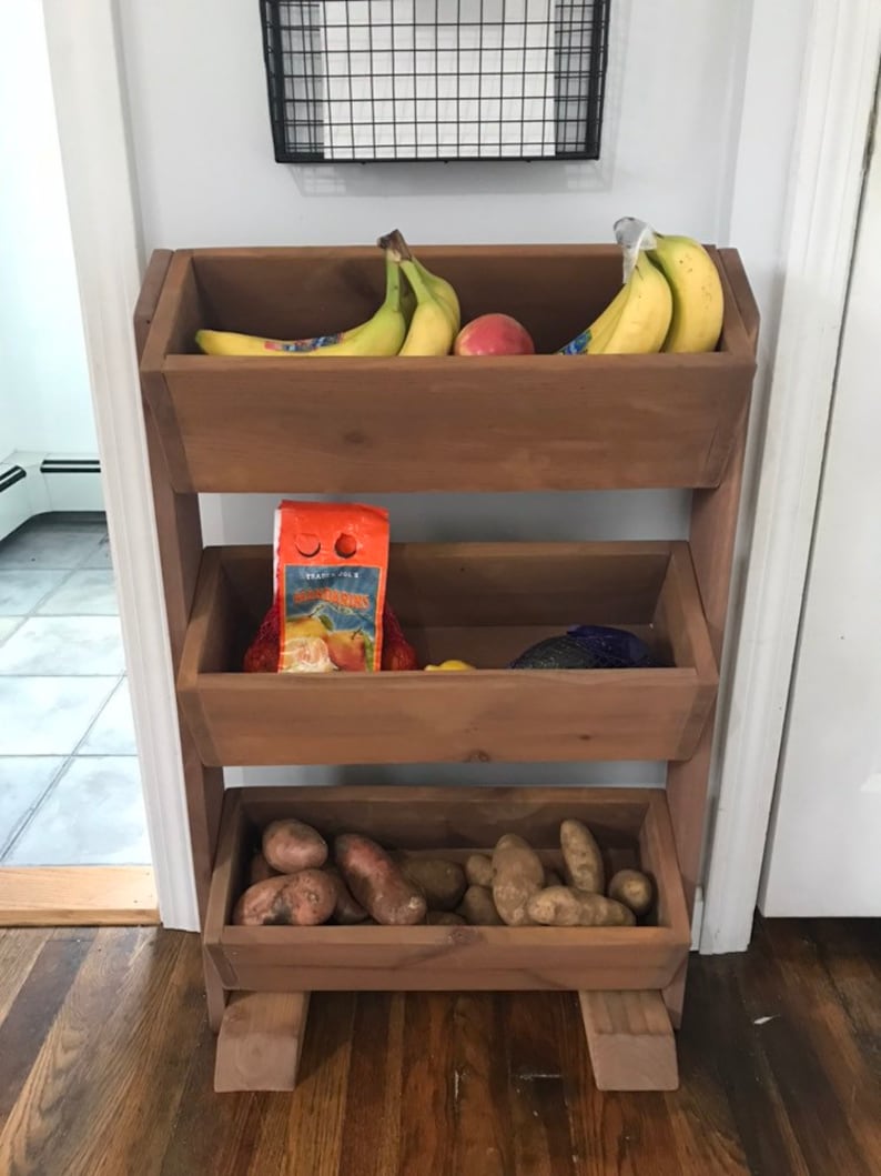 May include: A three-tiered wooden stand with three baskets. The top basket contains bananas and an apple. The middle basket contains a bag of orange slices. The bottom basket contains potatoes.