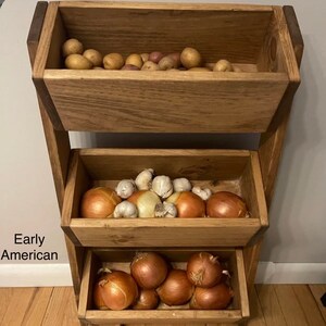 May include: A three-tiered wooden stand with three baskets, each filled with different vegetables. The top basket holds potatoes, the middle basket holds onions and garlic, and the bottom basket holds onions. The stand is made of natural wood and has a rustic look.