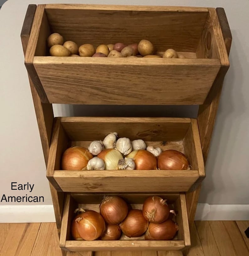 May include: A three-tiered wooden stand with three baskets, each filled with different vegetables. The top basket holds potatoes, the middle basket holds onions and garlic, and the bottom basket holds onions. The stand is made of natural wood and has a rustic look.