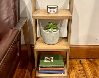 May include: A wooden shelf unit with three shelves. The top shelf has a candle with the text "HA" on it. The middle shelf has a small potted succulent plant. The bottom shelf has a stack of three books. The shelf unit is mounted to the wall.