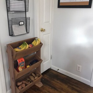 May include: A wooden tiered stand with four shelves, holding bananas, a bag of chips, and potatoes. The stand is in a kitchen setting with a white door and a black framed calendar on the wall.
