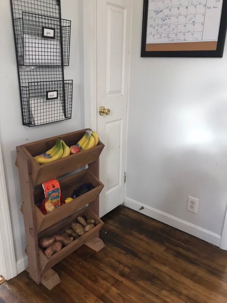 May include: A wooden tiered stand with four shelves, holding bananas, a bag of chips, and potatoes. The stand is in a kitchen setting with a white door and a black framed calendar on the wall.