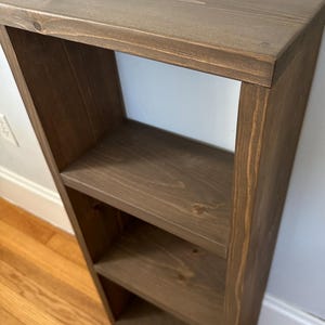 May include: A three-tiered wooden shelf unit with a dark brown stain. The shelf is made of solid wood and has a simple, rustic design.