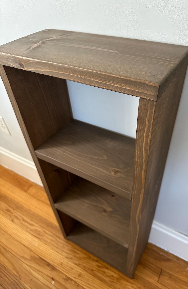 May include: A three-tiered wooden shelf unit with a dark brown stain. The shelf is made of solid wood and has a simple, rustic design.