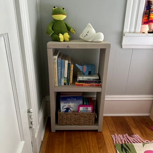 May include: A gray wooden bookshelf with two shelves. The top shelf has a green frog plush toy, a white cat plush toy, and a stack of books. The bottom shelf has a basket with a book titled "Frozen Snow Angels" and a pink tablet.