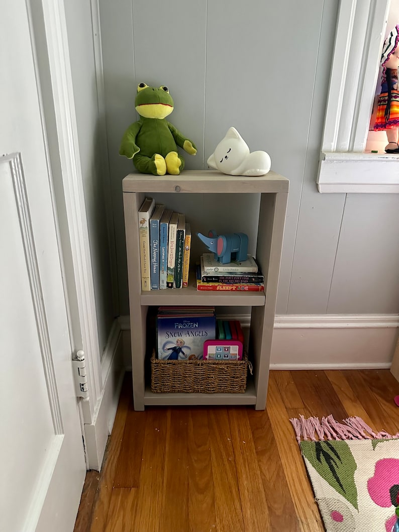 May include: A gray wooden bookshelf with two shelves. The top shelf has a green frog plush toy, a white cat plush toy, and a stack of books. The bottom shelf has a basket with a book titled "Frozen Snow Angels" and a pink tablet.