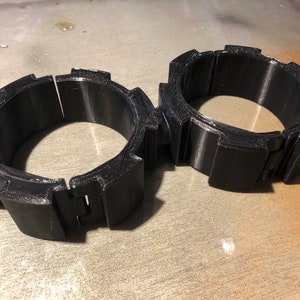 May include: Two black, segmented wrist cuffs. Each cuff is composed of several rectangular sections connected by hinges. The cuffs appear to be made of a hard plastic material. The design is angular and geometric.
