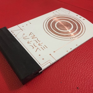 May include: A white rectangular object with a black edge and copper-colored circular and linear designs. The object has text that reads "FNIVE" with arrows. The item is on a red surface.