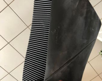 May include: A black radiator with horizontal metal fins and a black rubber cover. The radiator is held by a hand with a gold ring. The background is a tiled floor.