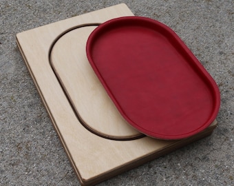 Puede incluir: Una tabla rectangular de madera clara con un recorte ovalado central, acompañada de una bandeja ovalada roja a juego. La bandeja está diseñada para encajar dentro del recorte de la tabla de madera, creando un conjunto con colores contrastantes.