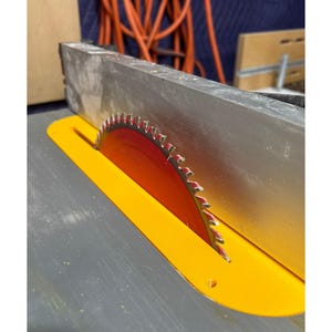 May include: Close-up of a table saw blade. The circular blade is red with silver teeth, set in a yellow safety guard. The saw's metal table is gray. The background includes orange electrical cords and a wooden structure.